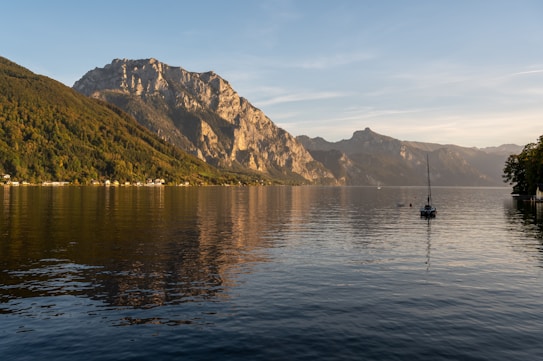 A serene landscape featuring a calm lake with gentle ripples, surrounded by mountains that capture the warm glow of the setting sun. A solitary sailboat rests on the water near the shore, with trees and small houses lining the base of the hills.