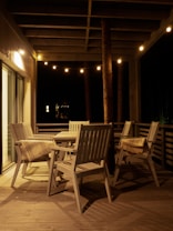 A cozy outdoor patio is illuminated by warm string lights. The space features wooden chairs and a table on a wooden deck, with a plaid blanket draped over one of the chairs. The setting suggests a serene and inviting atmosphere perfect for evening relaxation.