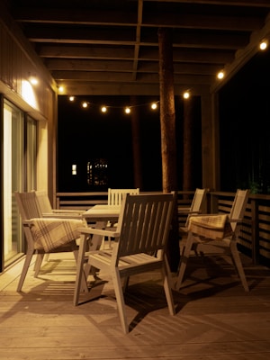 A cozy outdoor patio is illuminated by warm string lights. The space features wooden chairs and a table on a wooden deck, with a plaid blanket draped over one of the chairs. The setting suggests a serene and inviting atmosphere perfect for evening relaxation.