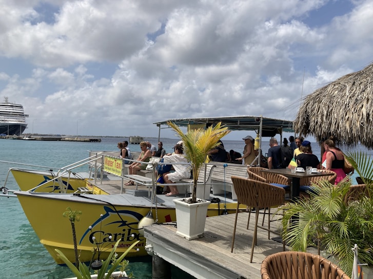 A group of people relaxing on a yellow water taxi docked at a pier. There are potted plants and wicker chairs on the dock, creating a casual and tropical atmosphere. In the background, a large cruise ship is visible against a partly cloudy sky.