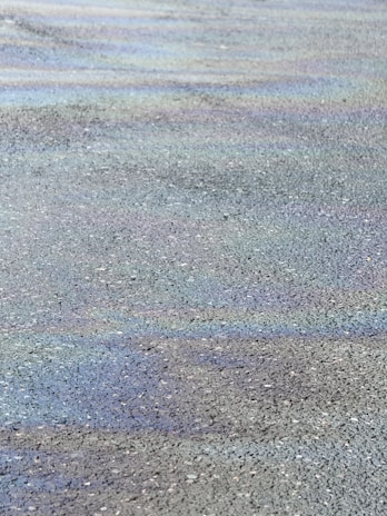 A view of an asphalt surface with visible rainbow-like oil slick patterns creating subtle iridescence. The texture of the road is coarse with small pebbles.