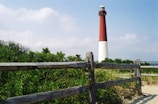 A tall red and white lighthouse stands prominently amidst green bushes and a wooden fence. The sky is partly cloudy with a natural, serene backdrop. The area surrounding the lighthouse appears to be sandy with coastal vegetation.
