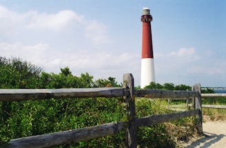 A tall red and white lighthouse stands prominently amidst green bushes and a wooden fence. The sky is partly cloudy with a natural, serene backdrop. The area surrounding the lighthouse appears to be sandy with coastal vegetation.