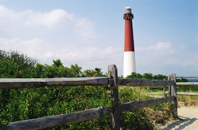 A tall red and white lighthouse stands prominently amidst green bushes and a wooden fence. The sky is partly cloudy with a natural, serene backdrop. The area surrounding the lighthouse appears to be sandy with coastal vegetation.