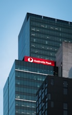 A modern high-rise building with sleek, reflective glass surfaces against a clear blue sky. The structure features a prominent red sign with white lettering that reads 'Australia Post'. The building's architecture includes dark rectangular sections and numerous well-lit office floors.