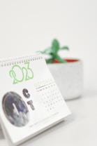 A desktop calendar displaying the month of October with the year 2023 prominently highlighted. The calendar has a spiral binding and is positioned on a white surface. In the background, a blurred green plant in a pot is visible, suggesting a minimalist and organized workspace.