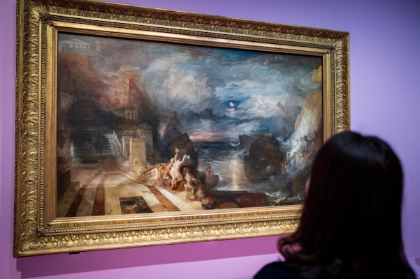 An ornate gold-framed painting hangs on a purple wall. The artwork depicts a historic or mythical scene featuring a dramatic landscape with rocky cliffs, clouds, and a stormy sea. Elegant architectural structures are visible, with figures interacting in the foreground.