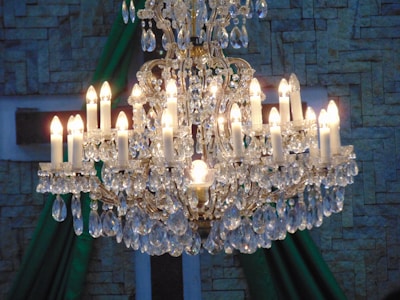 A luxurious chandelier with multiple lit candles and numerous crystal ornaments hanging in an elegant design. The chandelier is set against a textured wall which has a stone-like appearance with green drapery.