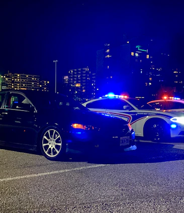 A nighttime scene showing a police car with flashing lights behind a pulled-over vehicle, driver visible through the windshield.