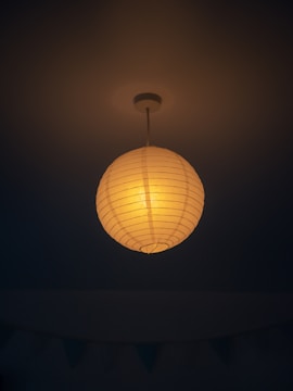 A warm, glowing paper lantern hangs from the ceiling, casting a gentle light against a dark background. The lantern is spherical with visible horizontal lines, suggesting a traditional paper crafting style.