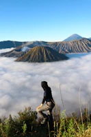 A traveler wearing rental hiking gear standing on a ridge overlooking an active volcano plume under a blue sky.