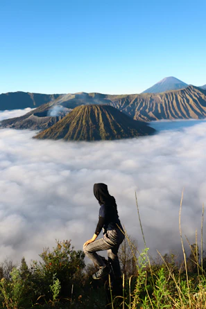 A traveler wearing rental hiking gear standing on a ridge overlooking an active volcano plume under a blue sky.