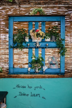 A vibrant setup featuring a bright blue wooden frame adorned with potted plants, including leafy greens and colorful flowers. Silver teapots and cups are elegantly positioned along the frame, while a decorative teapot and pitcher sit on the right. The background consists of a woven bamboo screen, providing a rustic touch. Below, text in Turkish is artistically handwritten on a turquoise wall.