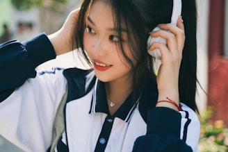 A person wearing a navy and white jacket listens to music on white headphones while smiling gently. The individual's long dark hair frames their face. The background suggests an outdoor setting with soft, blurred greenery.
