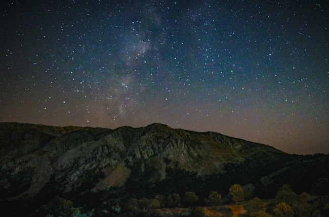 A clear, starry night sky with the Milky Way visible above a dark mountain range. The foreground has silhouetted hills and scattered trees, adding depth to the scene.
