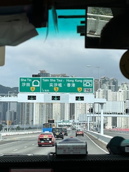 A busy highway scene with multiple vehicles including taxis and a truck moving towards the city. Overhead, a green road sign indicates directions to Sha Tin, Tsim Sha Tsui, and Hong Kong with distances and route numbers. Skyscrapers and tall buildings of the city are visible in the background under a cloudy sky.