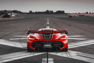 A vibrant red, high-performance sports car is positioned in the center of a deserted airstrip. The car is facing away from the viewer, showcasing its aerodynamic rear design and prominent spoiler. The surrounding landscape features blurred greenery and a cloudy sky, adding to the dramatic atmosphere.