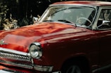 A classic vintage car with a shiny red exterior and chrome detailing is parked outdoors. Its curved design and polished surface reflect sunlight, highlighting the intricate craftsmanship. The front grille and headlights give a retro feel, with the steering wheel visible through the window.
