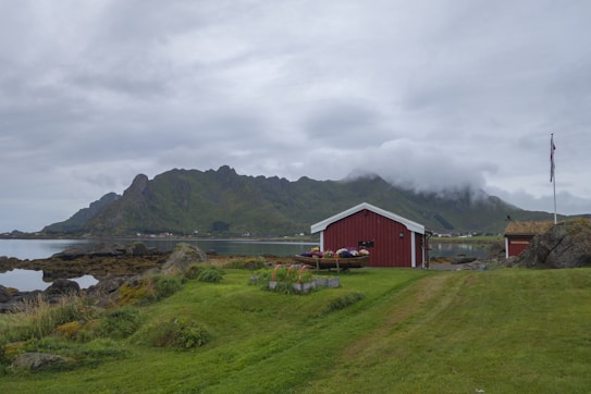 A red wooden cabin sits on a grassy area near the shoreline, with a mountainous landscape shrouded in clouds in the background. The foreground includes a neatly trimmed lawn and colorful flowers in a planter. A flagpole with a raised flag stands to the right.