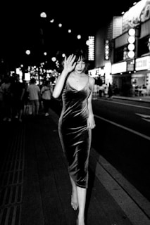 A woman in a velvet dress walks down a busy street at night, surrounded by bright lights and illuminated signs. She is holding her hand up near her face, walking confidently as other people can be seen in the background.
