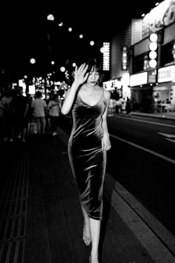 A woman in a velvet dress walks down a busy street at night, surrounded by bright lights and illuminated signs. She is holding her hand up near her face, walking confidently as other people can be seen in the background.
