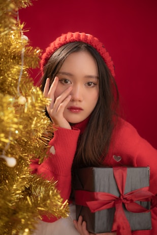 A young person wearing a red sweater and matching hat is holding a black gift box with a red ribbon. They are posing thoughtfully next to a decorated Christmas tree adorned with golden tinsel.