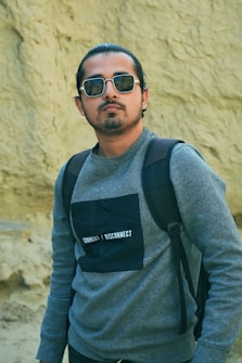 A person wearing sunglasses, a gray sweater with a black rectangle that says 'CONNECT | DISCONNECT,' and a backpack stands in front of a textured sandy-colored background.