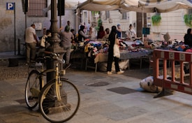 An outdoor market setup featuring multiple tables with assorted items for sale under white canopy tents. Various people are browsing and interacting with the vendors. A bicycle is parked against a lamppost in the foreground. The surrounding area includes some urban details such as a stone-paved floor, a barricade, a building with windows, and a parking sign.