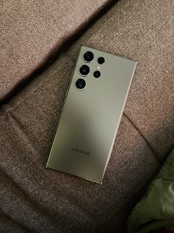 A sleek Samsung smartphone is placed on a brown fabric surface. The phone has a modern, minimalist design and features multiple camera lenses on its back. The fabric surface appears textured and soft.