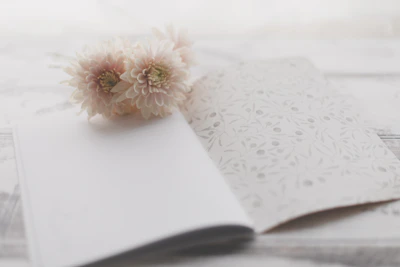 A soft-focus image showing a botanical-themed recipe card with gold accents resting on a cream linen cloth beside a small bowl of floral spread.