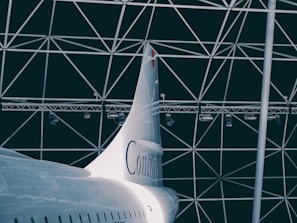 A sleek, white aircraft tail with the word 'Concorde' is positioned against a geometric, dome-like structure in the background. The metal beams criss-cross, forming triangular patterns. The lighting is dim, adding a dramatic flair to the scene.