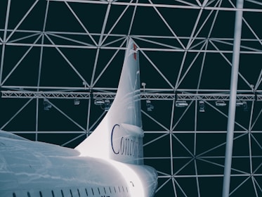 A sleek, white aircraft tail with the word 'Concorde' is positioned against a geometric, dome-like structure in the background. The metal beams criss-cross, forming triangular patterns. The lighting is dim, adding a dramatic flair to the scene.