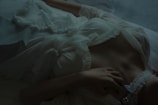 A person is lying down in a relaxed pose, wearing a white, ruffled garment with delicate lace and ribbon details. The lighting is dim and atmospheric, highlighting the textures of the fabric and the relaxed posture.