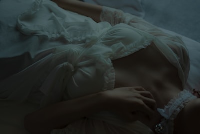 A person is lying down in a relaxed pose, wearing a white, ruffled garment with delicate lace and ribbon details. The lighting is dim and atmospheric, highlighting the textures of the fabric and the relaxed posture.