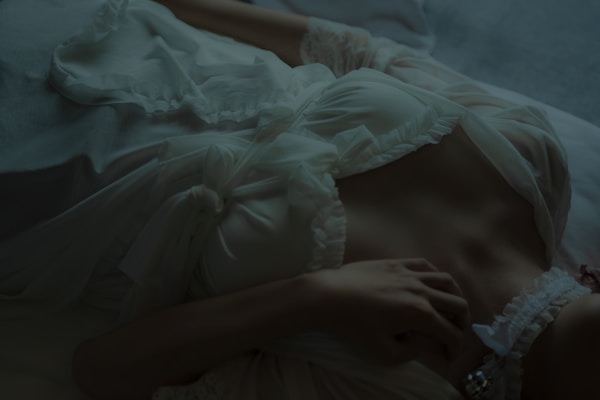 A person is lying down in a relaxed pose, wearing a white, ruffled garment with delicate lace and ribbon details. The lighting is dim and atmospheric, highlighting the textures of the fabric and the relaxed posture.