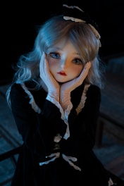 A doll-like figure with large, expressive eyes and long, blonde hair is posed with hands gently holding its face. The figure wears a dark, elegant dress with lace trim and a matching bow atop its head. The lighting casts a soft glow, emphasizing the delicate features and creating a mysterious ambiance.