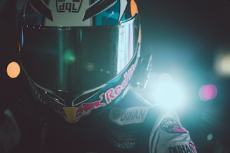 A motorcyclist wearing a helmet is positioned against a backdrop with bright lights. The helmet features a prominent visor and is adorned with various logos. The scene has a dramatic and intense lighting effect, primarily coming from one side.