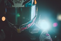 A motorcyclist wearing a helmet is positioned against a backdrop with bright lights. The helmet features a prominent visor and is adorned with various logos. The scene has a dramatic and intense lighting effect, primarily coming from one side.