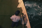 A person dressed in a uniform and cap stands on the deck of a ship next to coiled rope. The deck is green and the water below is dark with white-capped waves.