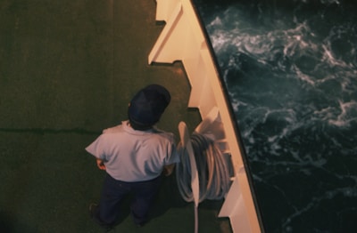 A person dressed in a uniform and cap stands on the deck of a ship next to coiled rope. The deck is green and the water below is dark with white-capped waves.
