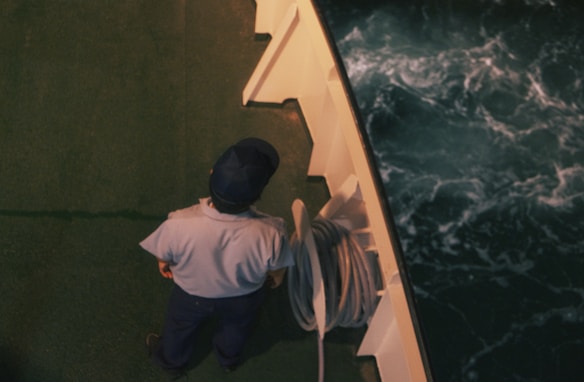 A person dressed in a uniform and cap stands on the deck of a ship next to coiled rope. The deck is green and the water below is dark with white-capped waves.
