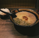 A black bowl of ramen with creamy broth, noodles, chopped green onions, and a dollop of red chili paste is situated on a wooden table. Beside it, there's a smaller bowl filled with white rice. The wooden table and the ambient lighting create a warm and inviting atmosphere.