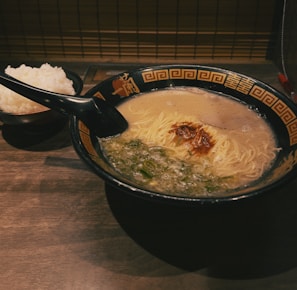 A black bowl of ramen with creamy broth, noodles, chopped green onions, and a dollop of red chili paste is situated on a wooden table. Beside it, there's a smaller bowl filled with white rice. The wooden table and the ambient lighting create a warm and inviting atmosphere.