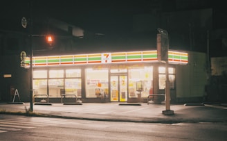 A brightly lit convenience store at night with a recognizable logo and signage glowing against the dark surroundings. The storefront is well illuminated, showcasing the various products inside. The empty parking lot and street in the foreground suggest a quiet, late-night setting.