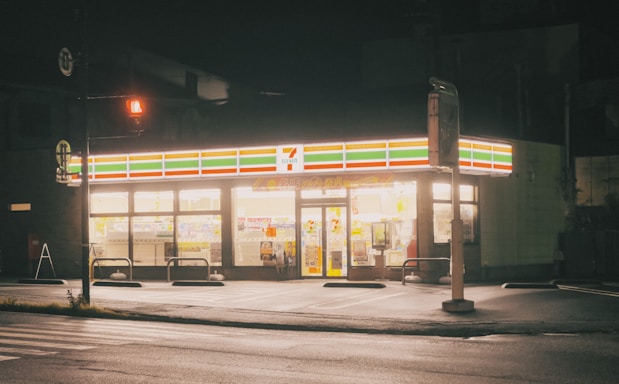A brightly lit convenience store at night with a recognizable logo and signage glowing against the dark surroundings. The storefront is well illuminated, showcasing the various products inside. The empty parking lot and street in the foreground suggest a quiet, late-night setting.