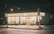A brightly lit convenience store at night with a recognizable logo and signage glowing against the dark surroundings. The storefront is well illuminated, showcasing the various products inside. The empty parking lot and street in the foreground suggest a quiet, late-night setting.