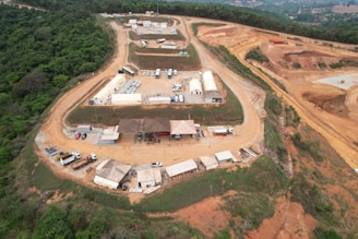 An aerial view of a construction or industrial site situated in a hilly and forested area. The image shows multiple white and beige temporary structures, likely tents or portable buildings, scattered across a large cleared area. There are several vehicles, including cars and trucks, parked near the structures. The surrounding landscape is a mix of greenery and cleared, reddish-brown dirt roads that wind through the site. The site appears to be segmented into different zones or sections.