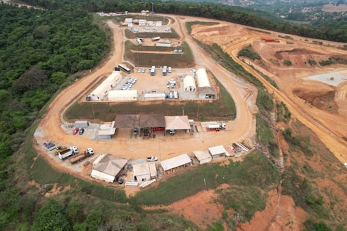 An aerial view of a construction or industrial site situated in a hilly and forested area. The image shows multiple white and beige temporary structures, likely tents or portable buildings, scattered across a large cleared area. There are several vehicles, including cars and trucks, parked near the structures. The surrounding landscape is a mix of greenery and cleared, reddish-brown dirt roads that wind through the site. The site appears to be segmented into different zones or sections.