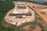 An aerial view of a construction or industrial site situated in a hilly and forested area. The image shows multiple white and beige temporary structures, likely tents or portable buildings, scattered across a large cleared area. There are several vehicles, including cars and trucks, parked near the structures. The surrounding landscape is a mix of greenery and cleared, reddish-brown dirt roads that wind through the site. The site appears to be segmented into different zones or sections.