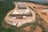 An aerial view of a construction or industrial site situated in a hilly and forested area. The image shows multiple white and beige temporary structures, likely tents or portable buildings, scattered across a large cleared area. There are several vehicles, including cars and trucks, parked near the structures. The surrounding landscape is a mix of greenery and cleared, reddish-brown dirt roads that wind through the site. The site appears to be segmented into different zones or sections.